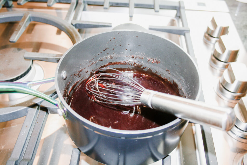 saucepan on a stove with red fruit gelatin mixture being stirred with a wire whisk