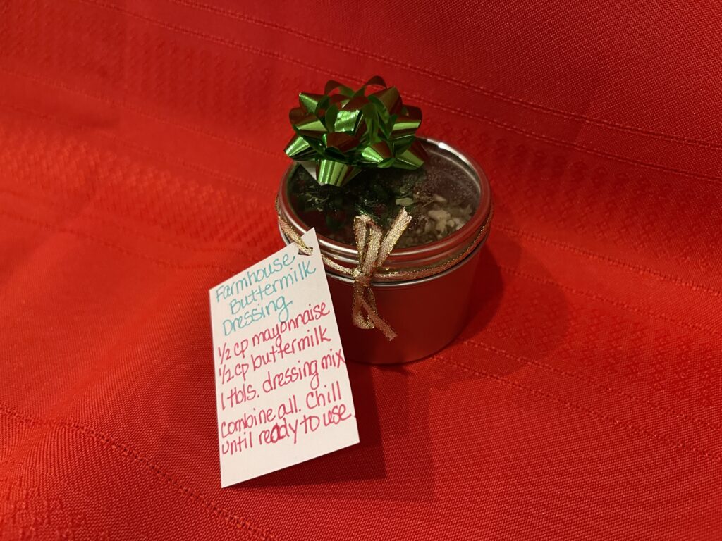 red background with small metal jar with clear, glass lid, containing dry ingredients for farmhouse buttermilk dressing mix. Green bow on top of container