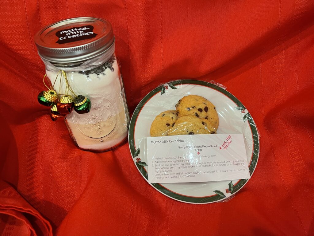 red background with a mason jar filled with dry ingredients for a batch of cookies. Jar is decorated with a label and some little bells and next to it is a christmas plate with 4 chocolate chip cookies and a recipe on top