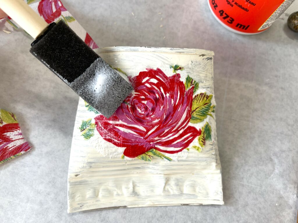 a foam paint brush decoupaging a rose paper napkin onto an ivory painted tin can flower holder