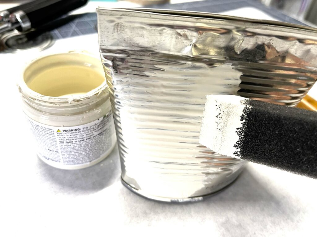 a tin can being painted with ivory colored paint and a sponge paint brush. A small jar of paint is sitting next to the can.
